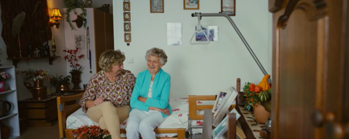 Persoon en oudere persoon zitten naast elkaar op een bed.